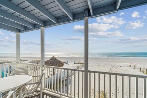 Ein überdachter Außenbereich mit Tisch und Stühlen mit Blick auf einen Strand und eine mit Reeds bedeckte Struktur.