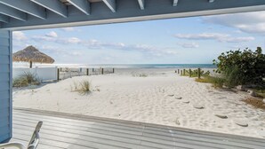 Une vue sur la plage depuis une terrasse couverte dotée d’un plancher en bois, une structure à toit de chaume, et une plage de sable avec une clôture et de la végétation.