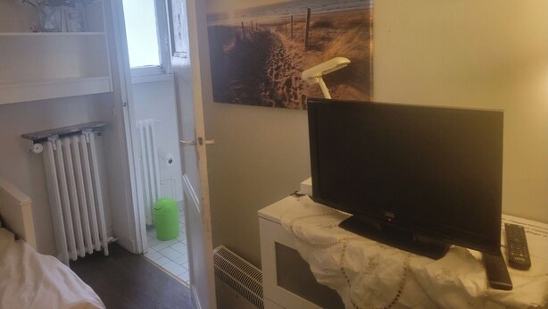 Une chambre d’hôtel avec un lit, un radiateur, une poubelle verte, un téléviseur posé sur un meuble et une horloge murale.