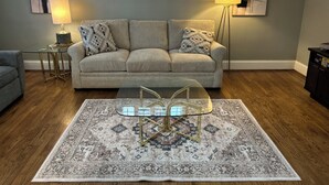 Ein Wohnzimmer mit einem beigen Sofa, einem gemusterten Teppich und einem Glastisch.