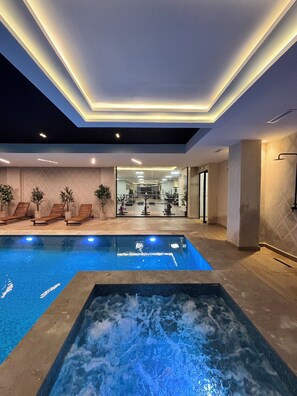 Een modern binnenzwembad met een jacuzzi, lounge stoelen en uitzicht op een fitnessruimte.