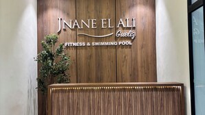 Ein Empfangstresen mit Holzoptik, ein Schild mit der Aufschrift 'JNANE EL ALI Fitness & Swimming Pool' und eine Topfpflanze auf der linken Seite.