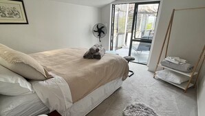 Une chambre avec un lit, un ventilateur sur pied, un tapis moelleux et un banc en bois.