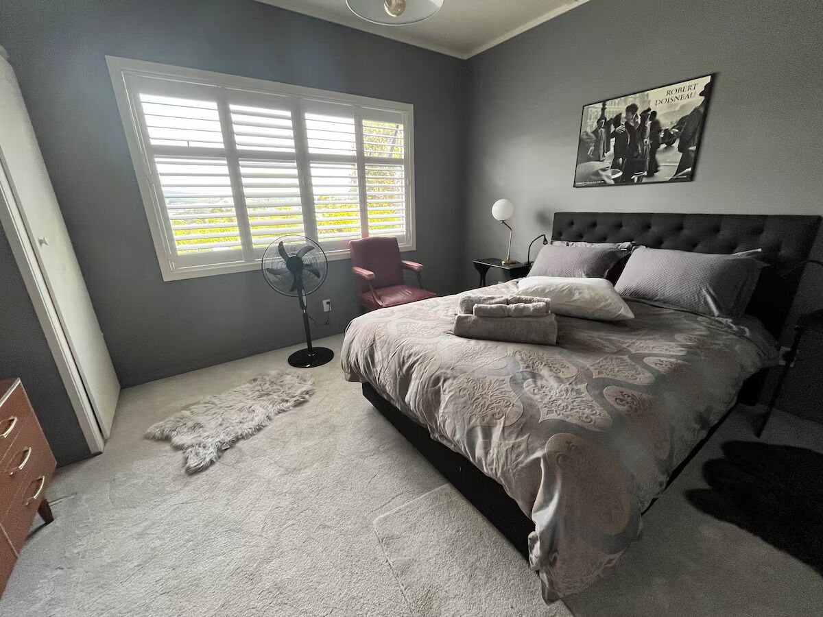 A bedroom with a bed, a chair, a standing fan, and a window with blinds.