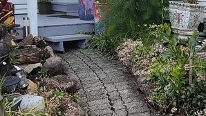 Un chemin mène à une maison blanche dotée d’un porche abrité, de plantes en pot et d’un petit jardin.