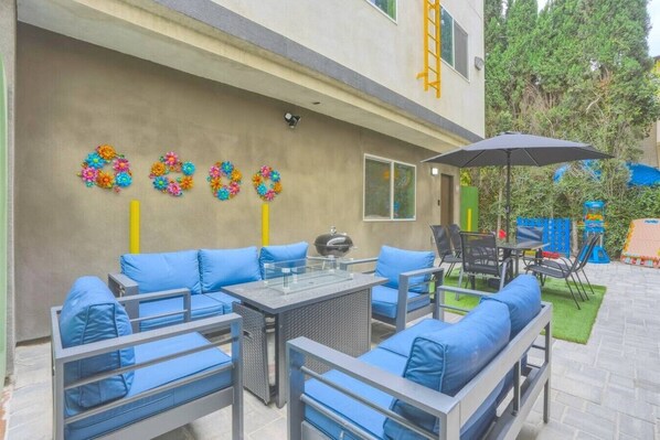 Un patio exterior con asientos acolchados azules, una mesa de vidrio y un paraguas negro.