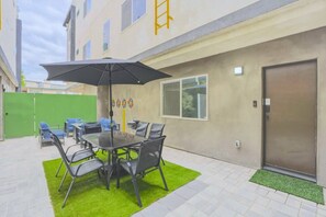 Un patio al aire libre con una mesa y sillas, un césped verde y un edificio con puerta y ventanas.