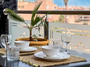A dining table set with white plates, bowls, and glasses, a potted plant, and a wooden bowl on a balcony with a view of buildings. - Superb Modern Studio in the heart of Guéliz - Sans Vis-à-Vis (Marrakech)