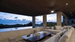 A modern living room with a large window overlooking a body of water and greenery. There is a sofa, a coffee table, and wicker ottomans. - Marea Surf Residences: 4BR, Private Pool & Golf Cart (Punta Mita)