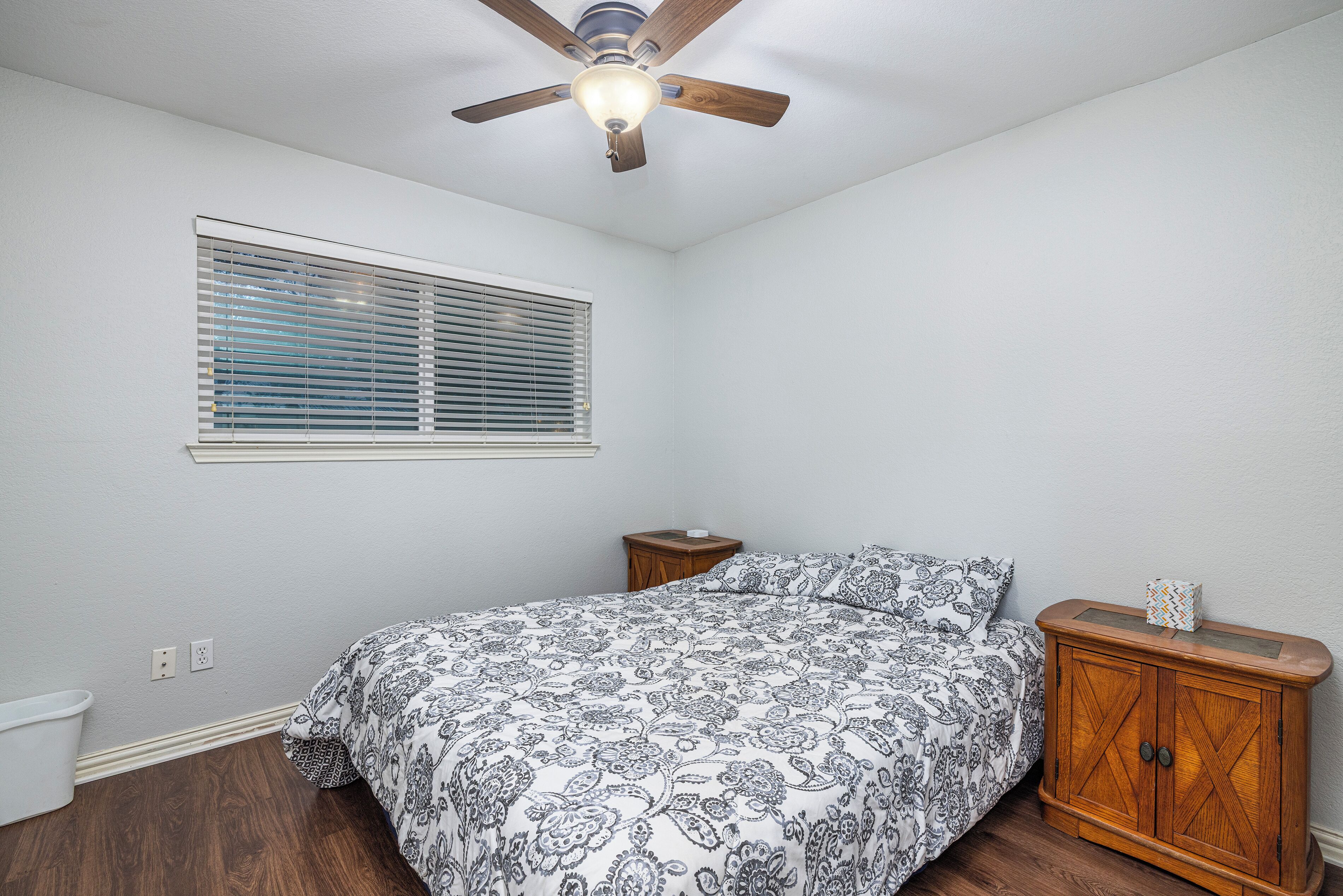 Une chambre à coucher avec un lit, une table de chevet en bois, un ventilateur de plafond et une fenêtre avec des stores.