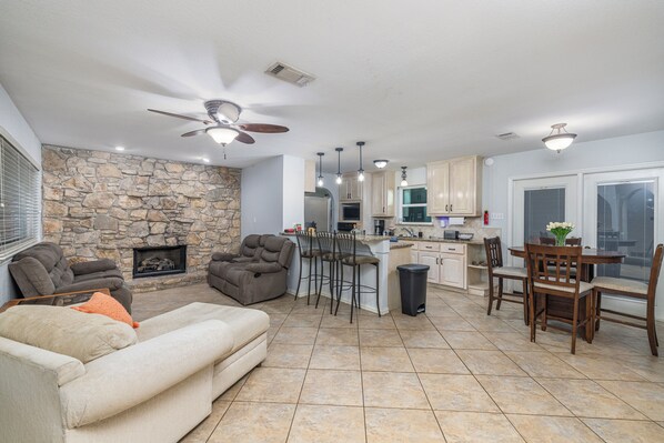 A spacious living area with a stone fireplace, a ceiling fan, and a kitchen with bar stools and a dining table. - Quiet, peacful, private 4-bedroom house with large backyard and patio space. (Arlington)