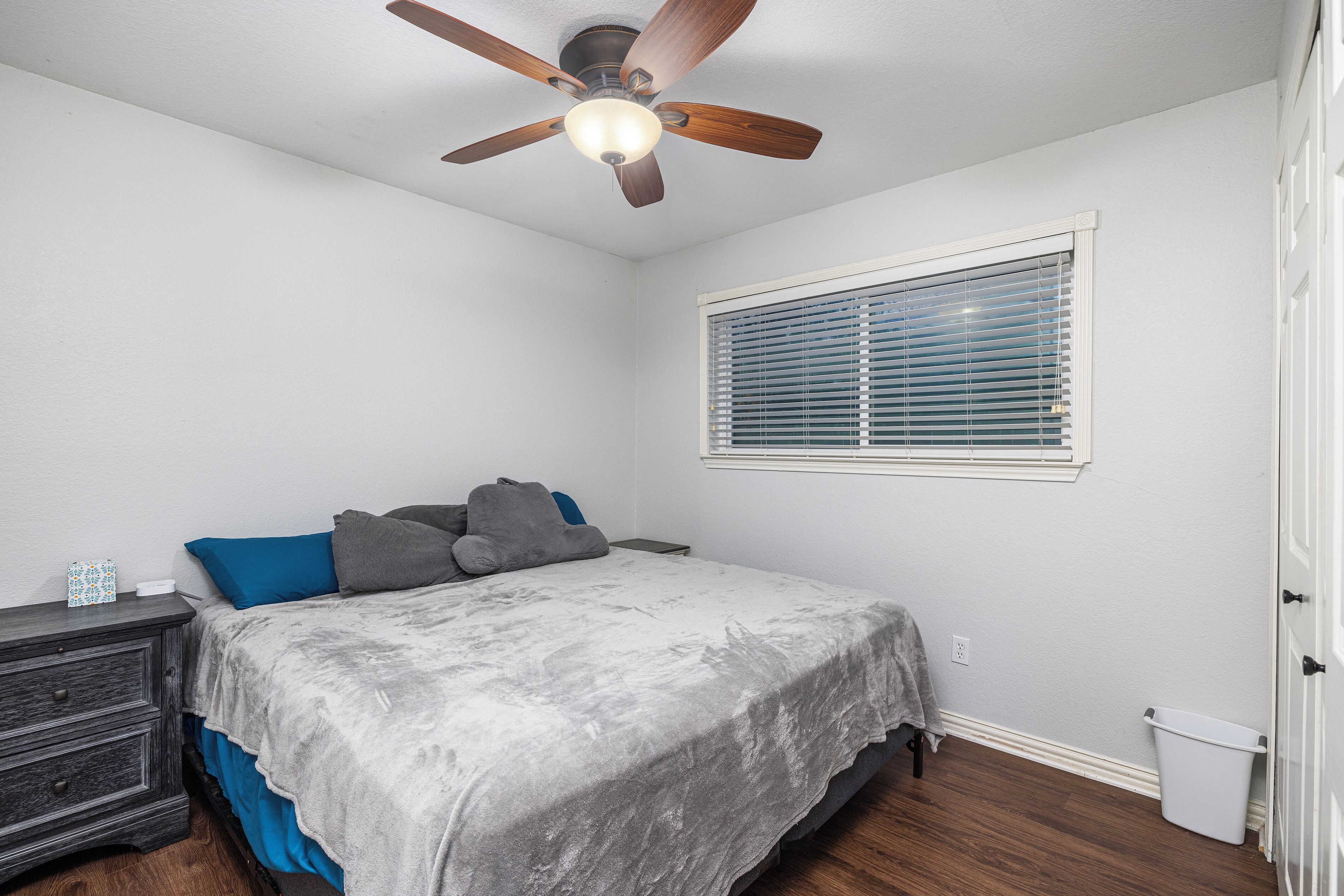 Une chambre à coucher comprenant un lit, une table de chevet, un ventilateur de plafond, une fenêtre avec des stores et une poubelle.
