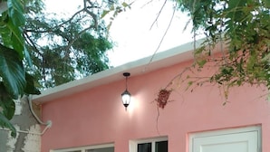 Um edifício cor-de-rosa com duas janelas e uma porta branca. Há duas cadeiras de metal preto e uma pequena mesa redonda.