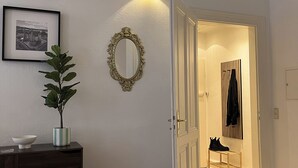 Käytävän lattia on puuta, siellä on tumma puinen kaappi laatikoineen, ruukussa oleva kasvi, peili ja kaksi kehystettyä valokuvaa seinällä.
