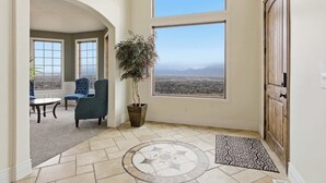 Amplia habitación con un ventanal que ofrece vistas a un paisaje urbano y montañas. Hay una planta en maceta y una silla azul cerca de la ventana.