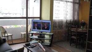 Een woonkamer met een televisie waarop twee personen te zien zijn, een eettafel met stoelen en een balkon met uitzicht op gebouwen.