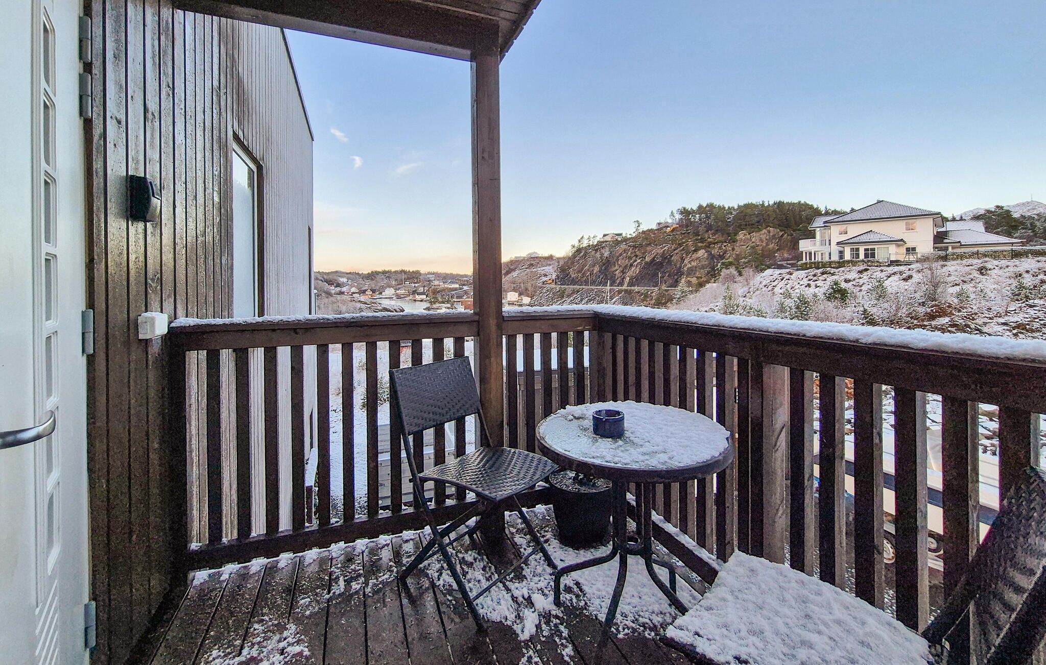 A wooden deck with a table and chairs, overlooking a snowy landscape and a building in the distance.