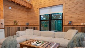 Un intérieur chaleureux avec des éléments en bois, notamment un canapé d’angle beige, une table basse en bois sur laquelle sont posés des livres, et une fenêtre équipée de stores.