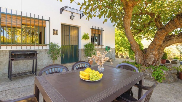 An outdoor dining area with a table set for two, surrounded by chairs, a grill, and a tree providing shade. - El Moral Prado del Rey by Ruralidays (Cádiz)