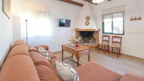A living room with a sofa, a wooden table, and chairs. There is a fireplace with a TV above it. - El Moral Prado del Rey by Ruralidays (Cádiz)