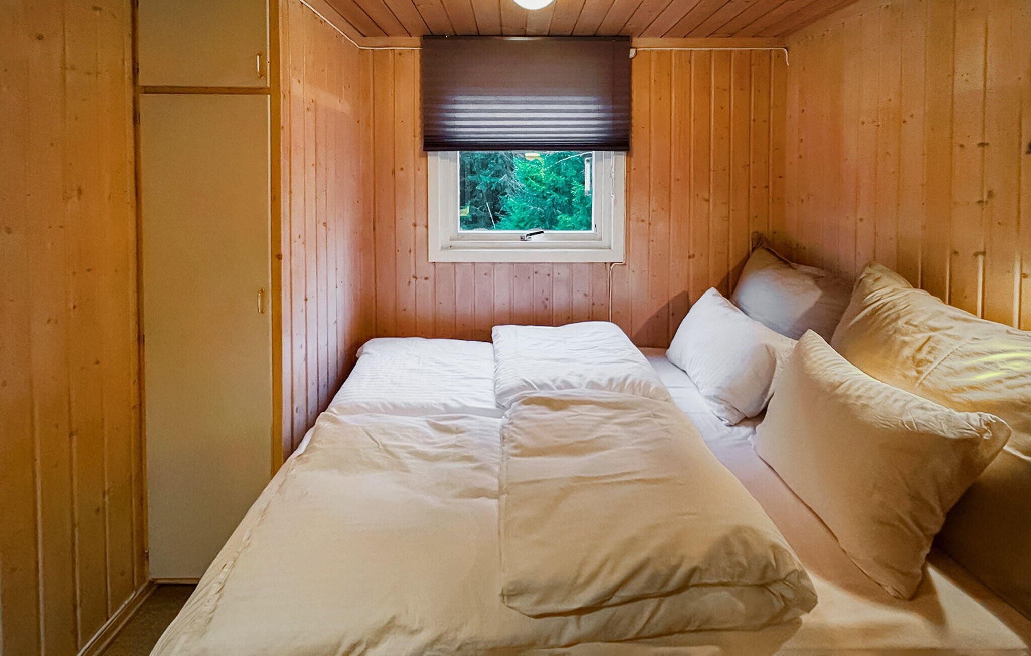 A small, cozy room with wooden walls, a single bed with white linens, a window with a view of greenery, and a wooden wardrobe.