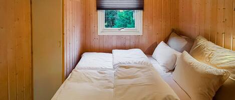 A small, cozy room with wooden walls, a single bed with white linens, a window with a view of greenery, and a wooden wardrobe.