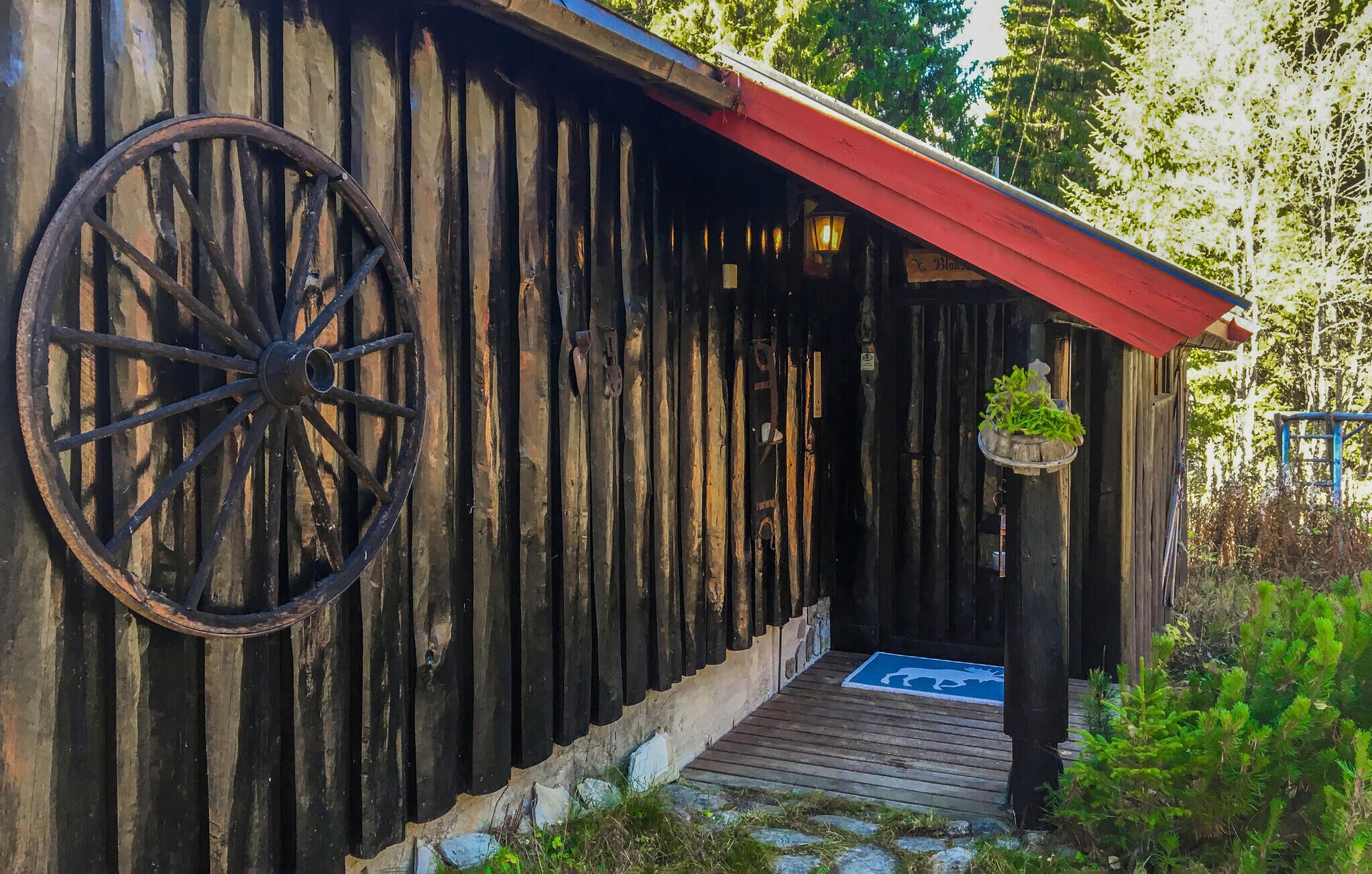 En rustik träbyggnad med ett stort hjul fäst på väggen, ett rött tak och en träveranda som leder till ingången.