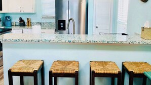 A modern kitchen with white cabinets, a stainless steel refrigerator, a coffee maker, and a bar area with stools. - Saltwater Solace, For 8-FREE WIFI/FREE PARKING/FREE BEACH ACCESS (Galveston)