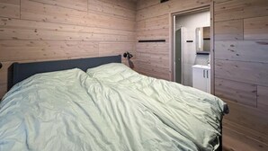 Ein Schlafzimmer mit Holzboden, einem Bett mit grauem Kopfteil, einer Nachttischlampe und einer Tür zu einem Badezimmer mit Spiegel und Waschbecken.