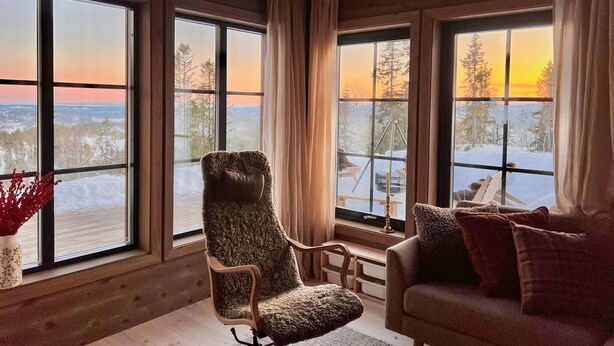 Ett mysigt rum med stora fönster som bjuder på en utsikt över ett snötäckt landskap och en solnedgång.