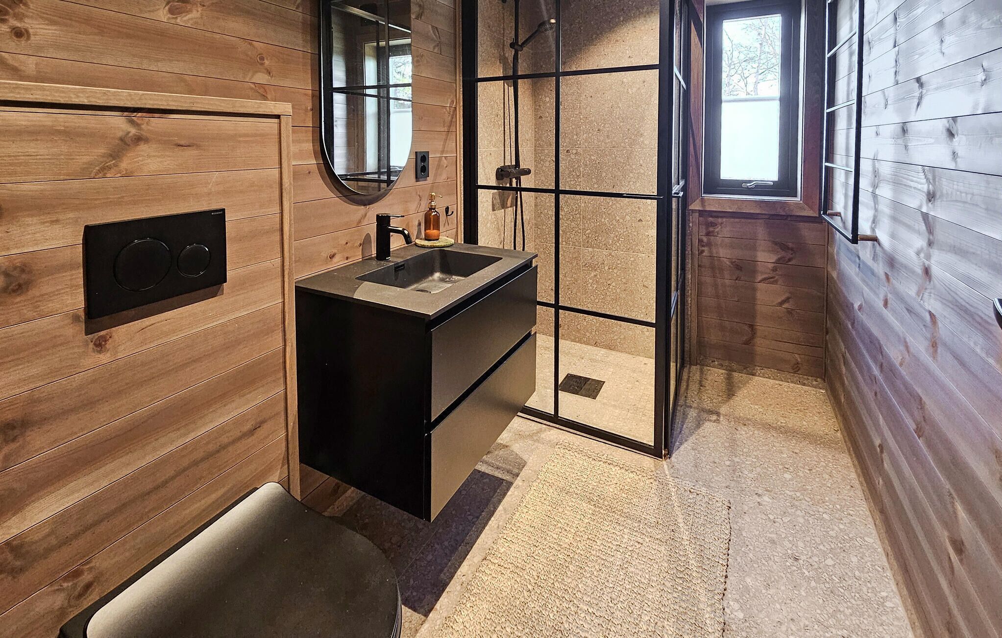 A bathroom with wooden walls, a sink with a mirror, a shower, and a window.