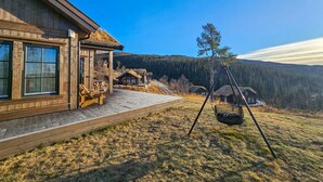 En stuga av trä med en veranda, en gunga och utsikt över ett bergigt område täckt av skog.