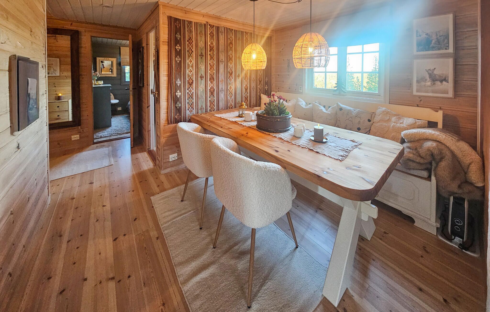 Un comedor de madera con una mesa larga, sillas blancas y un banco. Hay luces colgantes y una ventana con obras de arte en la pared.