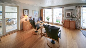 Ett modernt vardagsrum med trägolv, en soffa, ett soffbord, en tv och ett skåp. Det finns stora fönster med gardiner och utsikt över omgivningen.