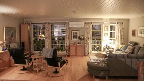Ett vardagsrum med en soffa, två fåtöljer, ett litet bord och ett skåp. Det finns ett fönster med gardiner och utsikt över omgivningen.