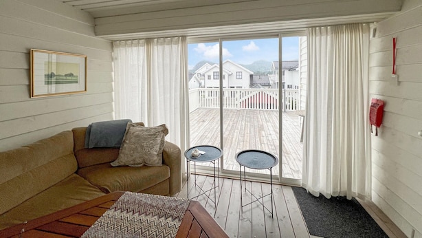Ett mysigt rum med trägolv, en beige soffa, två små, runda bord och utsikt över hus genom ett stort fönster med vita gardiner.