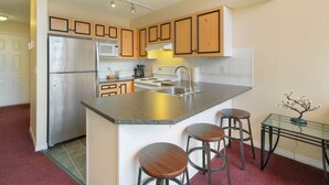 Een moderne keuken met een roestvrijstalen koelkast, magnetron en fornuis, houten kastjes, een granieten kookeiland met krukken en een kleine tafel met een decoratieve plant.