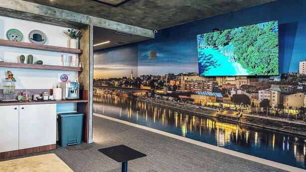 A modern bar area with a large TV screen displaying a cityscape, a counter with various items, and a view of a river. - Circel of 2 (Belgrade)