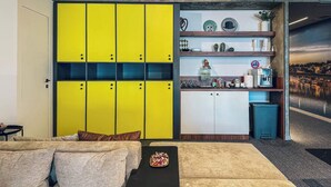 A modern living room with a sofa, a coffee table with a pizza, and a kitchen area with yellow cabinets and various kitchen items. - Circel of 2 (Belgrade)