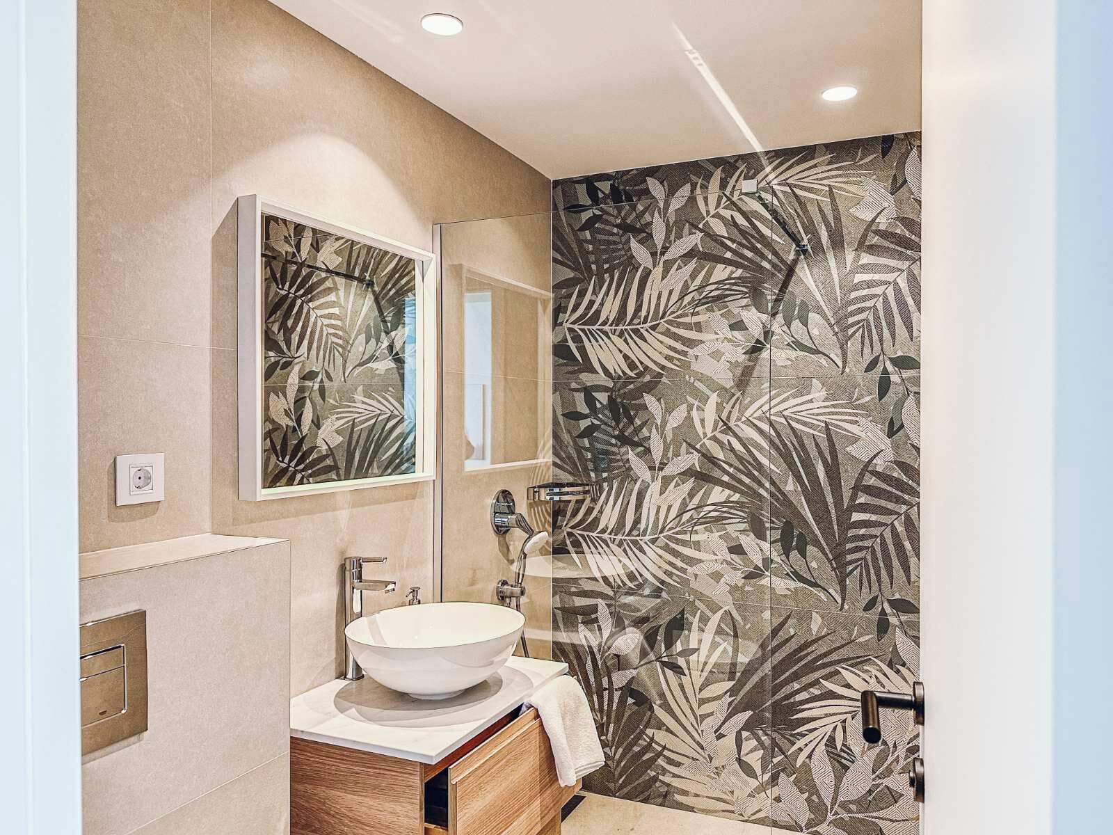 A modern bathroom with a large mirror, a white sink, and a shower with a leaf-patterned wall.
