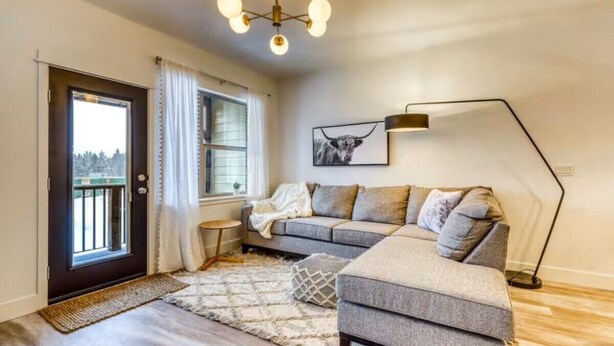Un salon moderne avec un canapé d’angle gris, un lampadaire et une grande fen être agrémentée de rideaux.