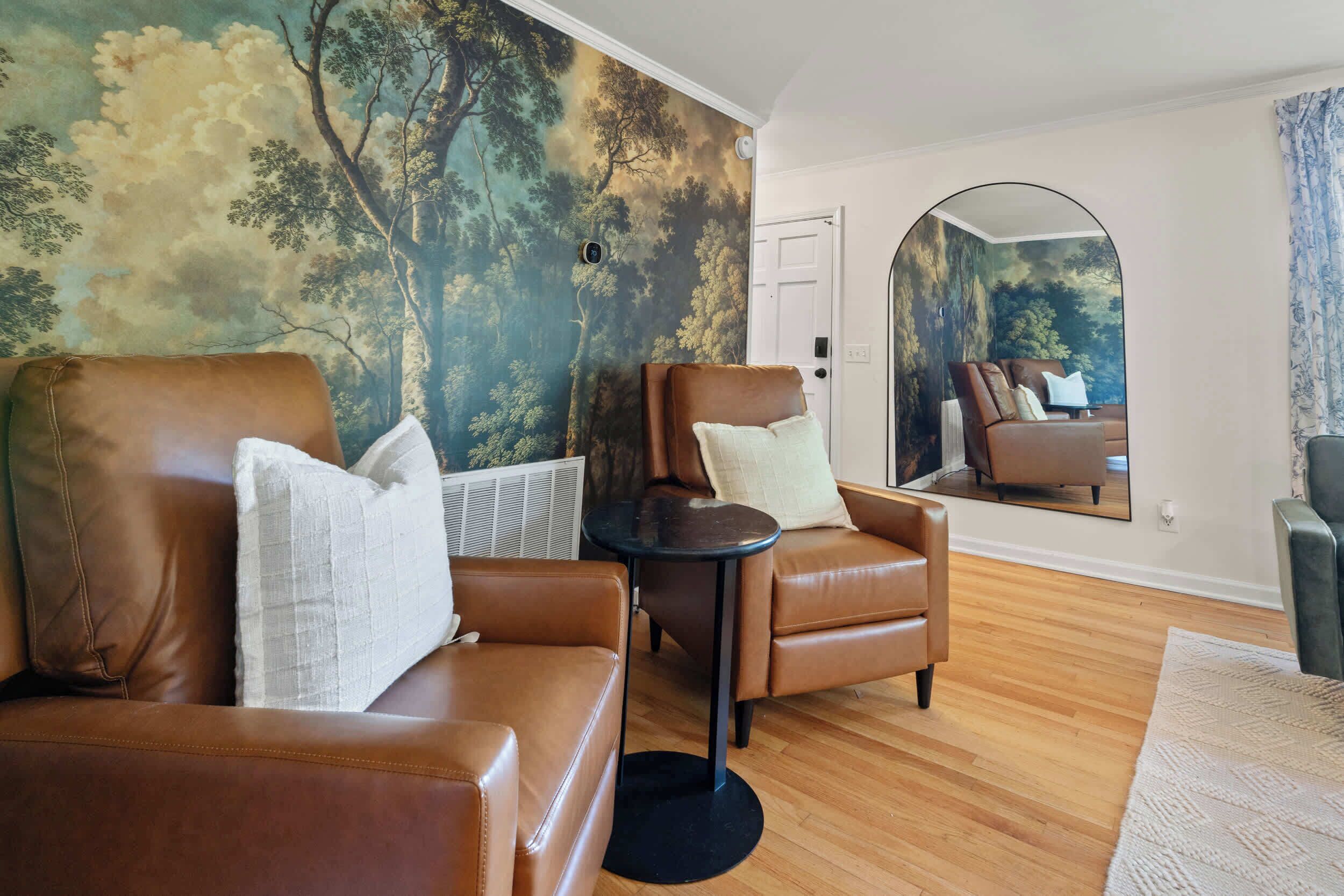 A room with a brown leather sofa, a matching armchair, and a small round table. A large mirror reflects the room, showing a door and a window with curtains.