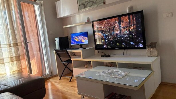 A modern living room with a large flat-screen TV, a sofa, a glass coffee table, and a sliding glass door. - Studio Dunja (Niksic)
