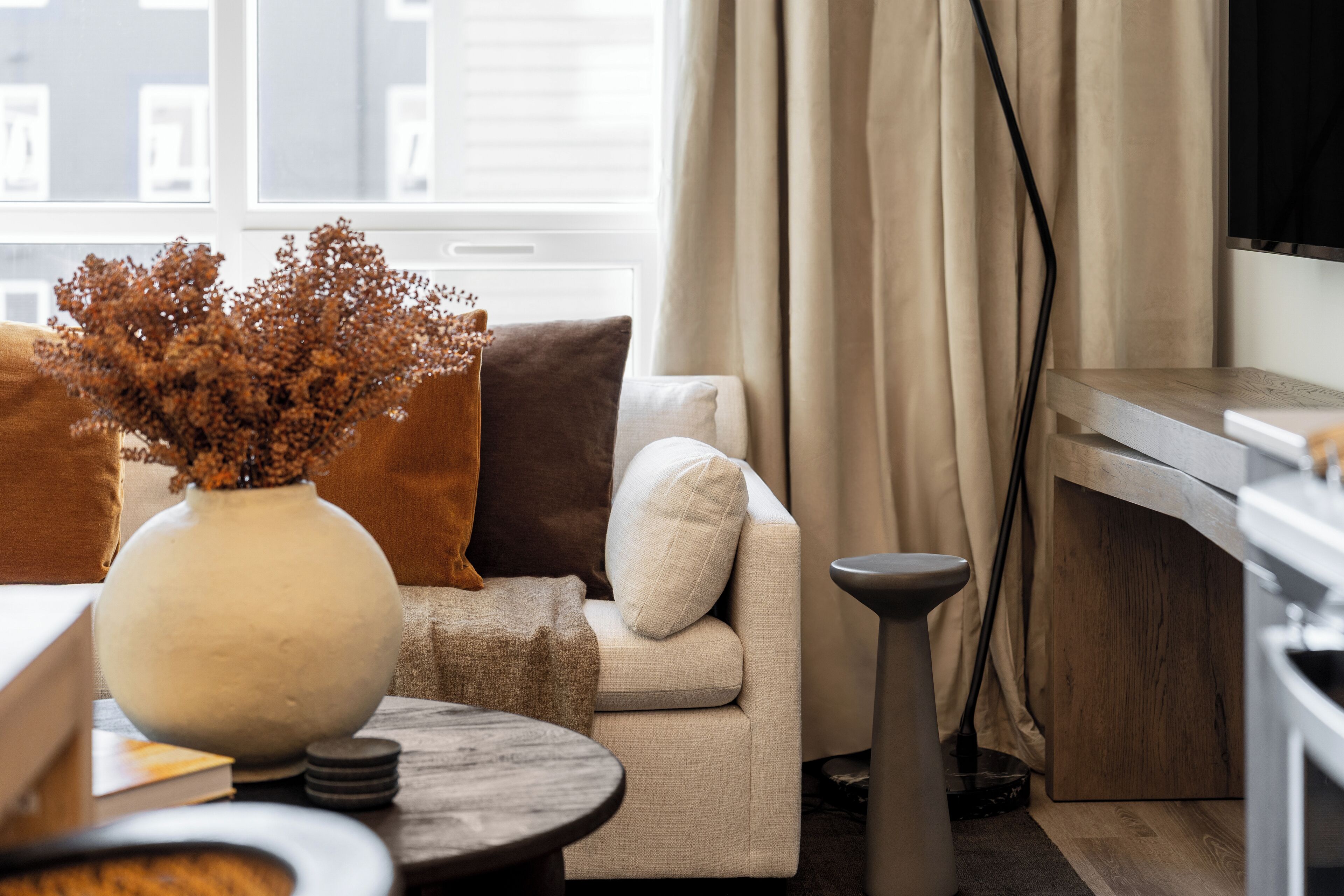 A cozy living room with a sofa, a round table, a vase with dried flowers, and a view of the city through a window.