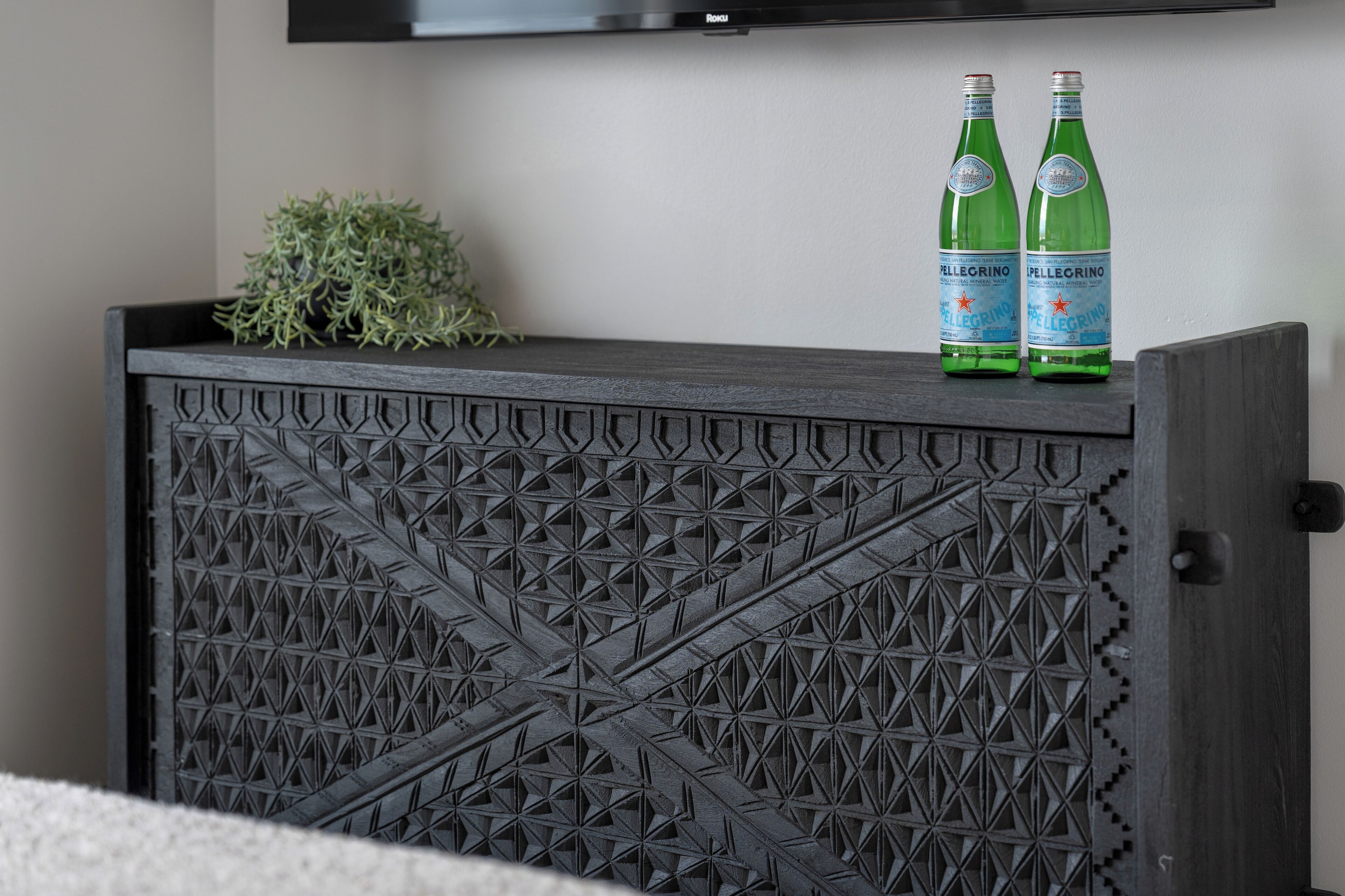 Un mobiletto in legno scuro con motivo geometrico, due bottiglie di acqua minerale Pellegrino e una piccola pianta sopra.