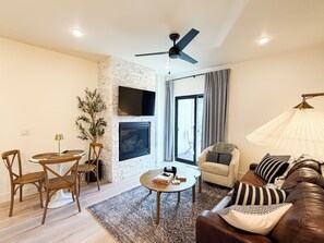 Ein Wohnzimmer mit Kamin, Deckenventilator, Wand-Fernseher, einer Couch, einem Couchtisch und einem Essbereich mit Stühlen und Tisch.