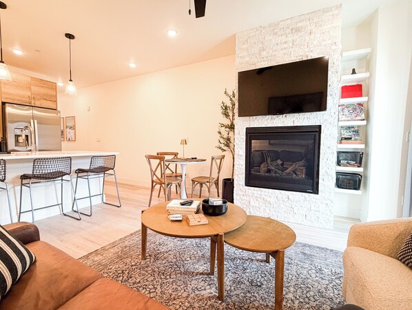 Ein modernes Wohnzimmer mit Kamin, einer Küchenzeile inklusive Kühlschrank und einem Essbereich mit Stühlen und Tisch.
