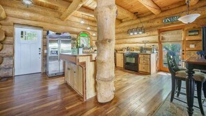 Interior de una cabaña de troncos con techo de madera, una zona de cocina, una mesa de comedor y un gran pilar de madera.