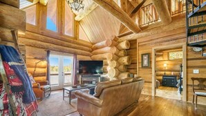 Interior de una cabaña de troncos con chimenea, sofá de cuero, piso de madera y televisor.