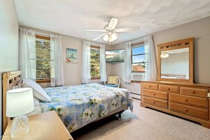 Un dormitorio con cama, mesita de noche, cómoda, ventilador de techo y ventana con cortinas.